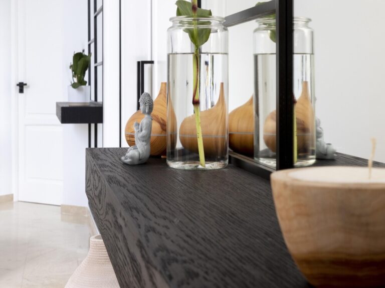 Detalle de interiorismo de lujo silencioso con una estantería de madera oscura de veta marcada, decorada con un jarrón de cristal, una figura zen y elementos naturales en un ambiente minimalista y luminoso.