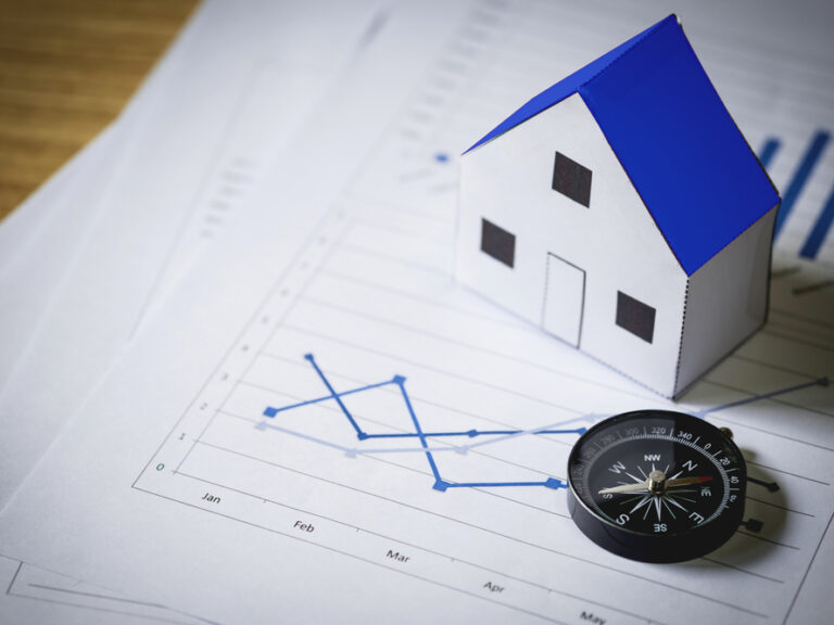 Una pequeña maqueta de una casa de papel con tejado azul situada sobre gráficos estadísticos financieros y una brújula, representando el análisis y la dirección del mercado inmobiliario.