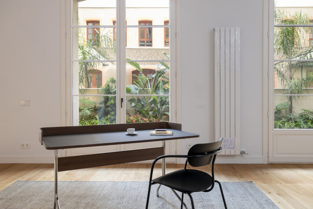 Una sala de estar moderna y luminosa que ofrece confort térmico en arquitectura gracias a los grandes ventanales que dan a un frondoso patio y un radiador vertical en la pared. Una mesa de escritorio negra con una taza de café y un libro está situada en primer plano, flanqueada por una silla de diseño negra.