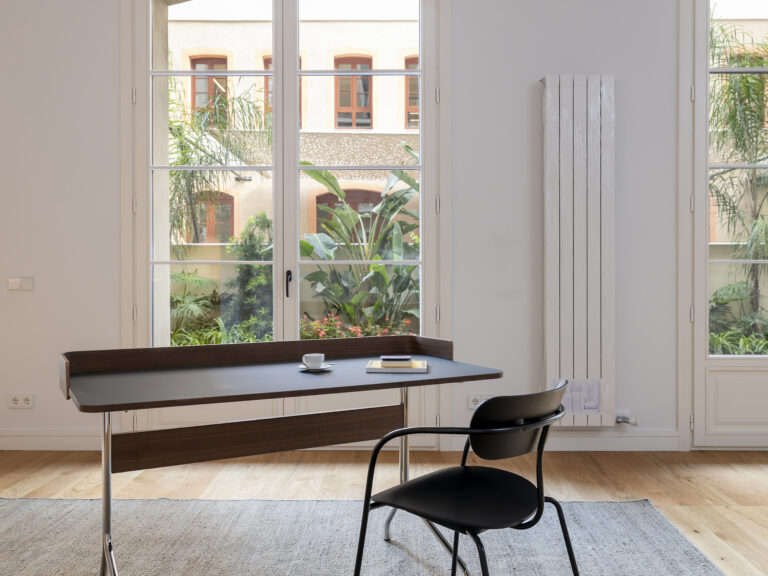 Una sala de estar moderna y luminosa que ofrece confort térmico en arquitectura gracias a los grandes ventanales que dan a un frondoso patio y un radiador vertical en la pared. Una mesa de escritorio negra con una taza de café y un libro está situada en primer plano, flanqueada por una silla de diseño negra.