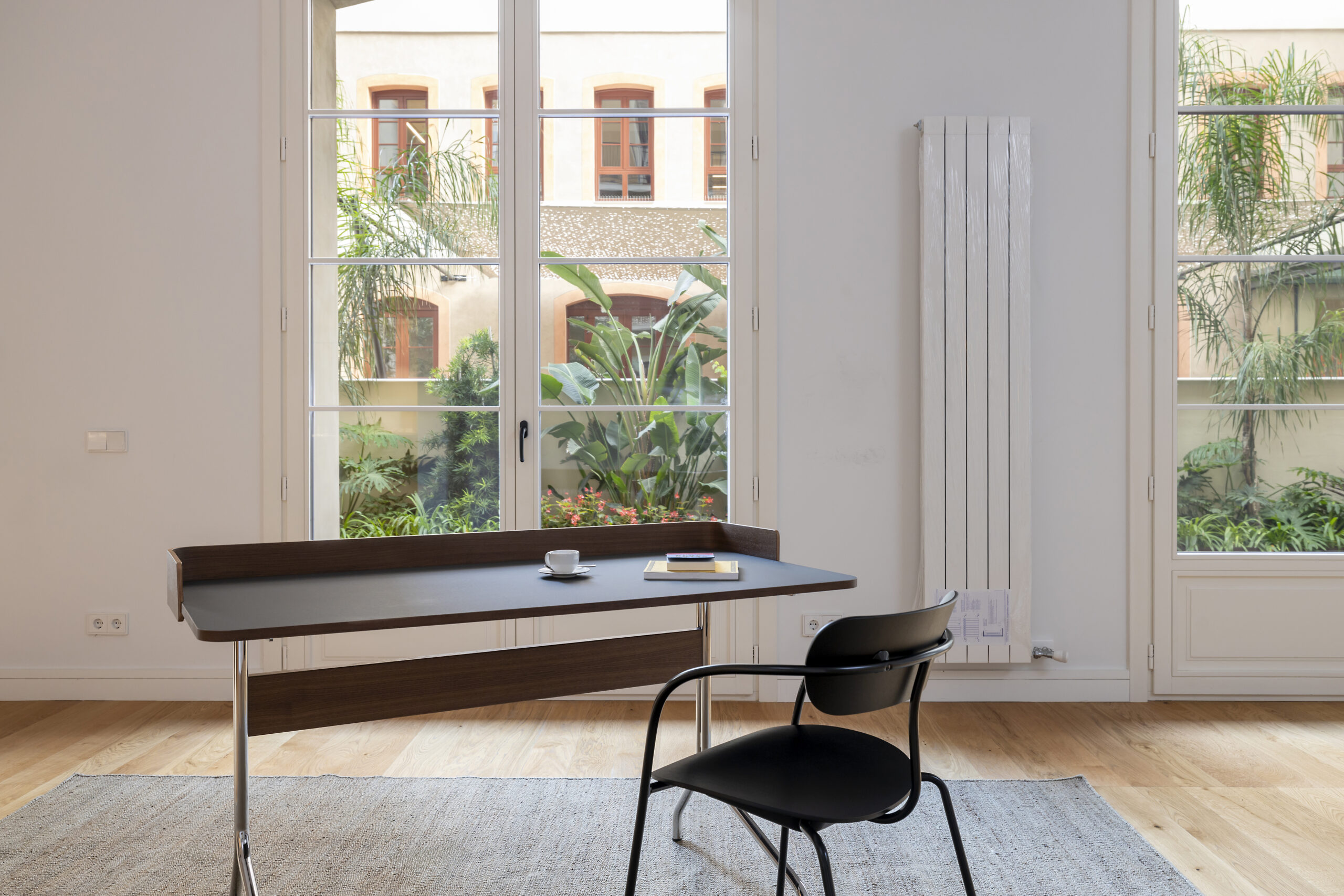 Una sala de estar moderna y luminosa que ofrece confort térmico en arquitectura gracias a los grandes ventanales que dan a un frondoso patio y un radiador vertical en la pared. Una mesa de escritorio negra con una taza de café y un libro está situada en primer plano, flanqueada por una silla de diseño negra.